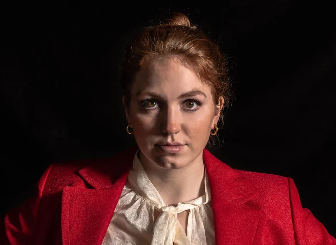 Professionally dressed woman in red blazer and white blouse looks straight into camera against black background
