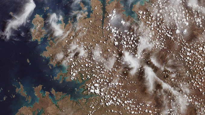  A satellite view of earth from space. Ocean is to the far left of the image and then brown, dry looking land area with puffy white clouds make up the rest of the view. 