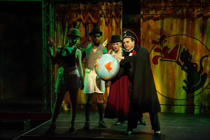 Actors stand on a stage in costume, looking at a globe one of them is holding in their hands.