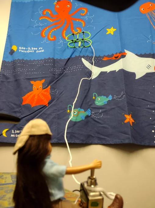 Lottie doll is seated and ready to drive her underwater robot near an octopus pictured in the wall hanging. The octopus lives in the Twilight zone in this ocean image, so the underwater robot will allow Lottie to explore the octopus habitat even though this part of the ocean is usually beyond diver depth. 
