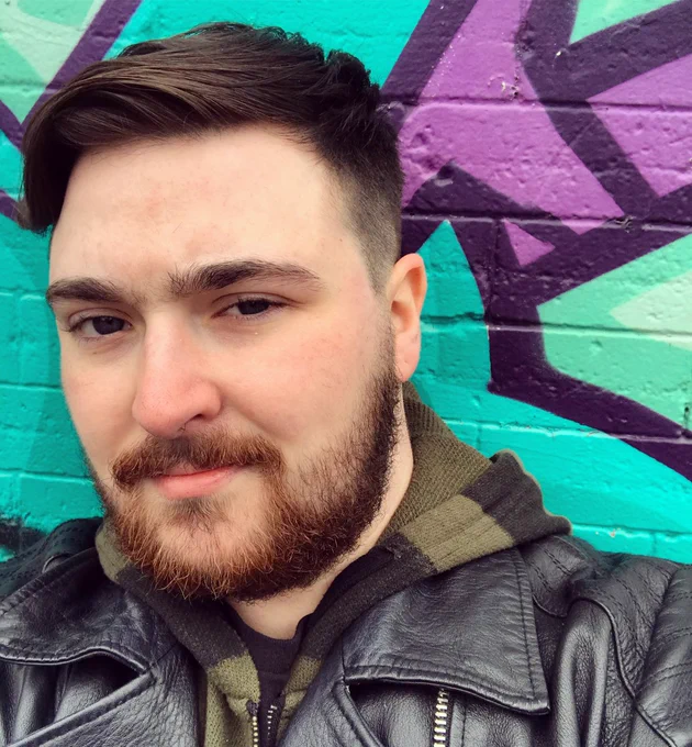 Asher, a man with short brown hair and a trimmed beard, wears a knit green and black striped sweater under a black leather jacket. He is standing in front of a purple, blue, and blue-green graffiti wall. 