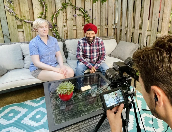 Elena Hudgins Lyle and Harvinder Wadhwa are smiling and sitting together on an outdoor couch. They are being filmed and can be seen on the viewfinder of the camera as well.