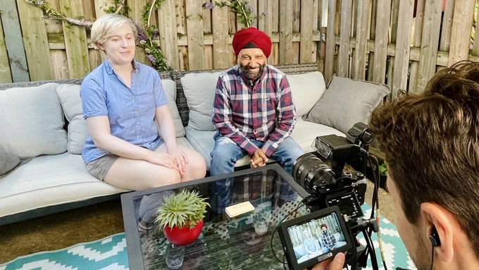 Podcast hosts Elena and Harv sit on a couch outdoors. Elena is wearing a blue button-up short sleeve shirt and has short blond hair; Harv is wearing a red turban and a long-sleeve plaid button-up.