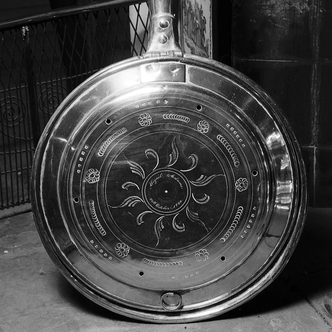 Black and white photo of a circular metal pan with a long wooden handle. Abigail Adams and 25th October 1764 are inscribed in the center. Embellishments are engraved on the pan in circular and leaf like patterns.