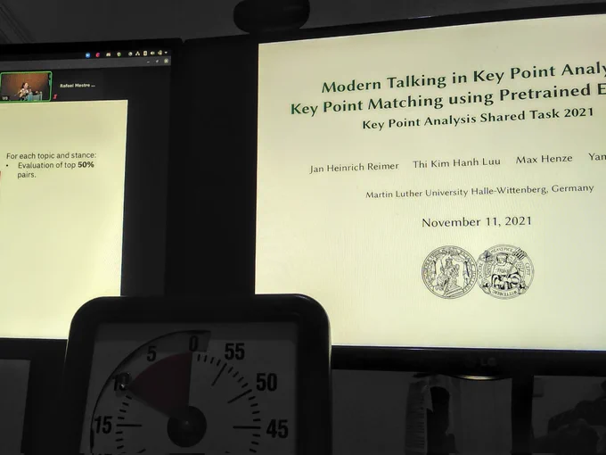 Two computer screens showing the Key Point Analysis shared task presentation Zoom conference, and a timer clock.