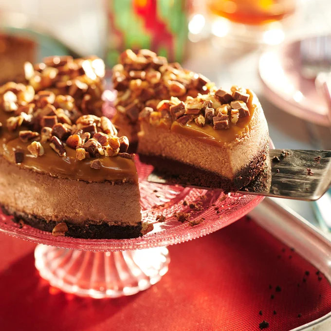 someone taking a slice from a caramel, chocolate and pecan-topped cheesecake