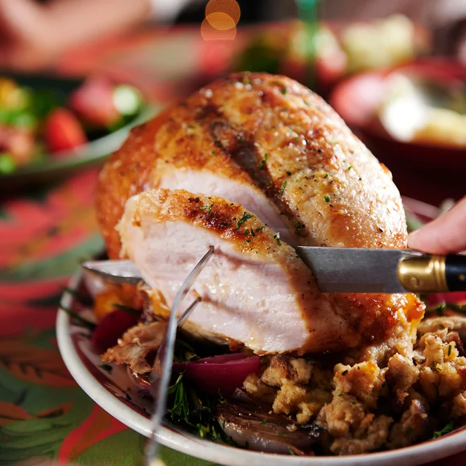 a carving fork and knife are slicing a piece off of a turkey breast, on a plate with stuffing and roasted vegetables. 