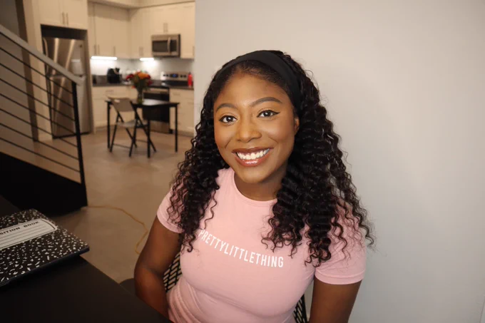 Girl smiling with a pink shirt on. In the background her kitchen is to her left and a blank wall to her right. She has a laptop on front of her