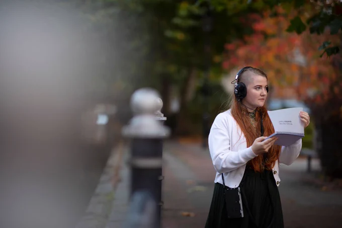 An image of a girl with long hair, wearing a white cardigan and black headphones. She's holding a guidebook and looking over in the distance. 