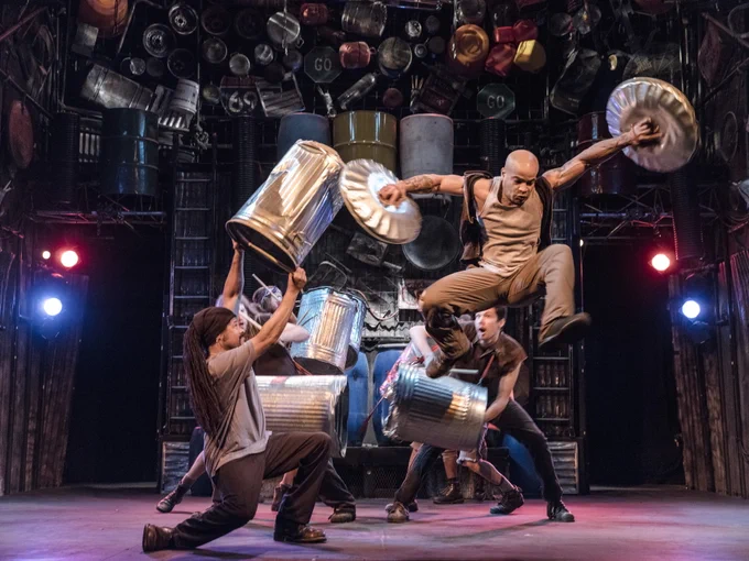 Five performers holding trashcans with one performer jumping and hold two trash can lids