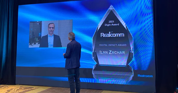 Live event photo from the Realcomm Digie Awards. Ilan Zachar from Carr Properties stands on stage in a blue business suit with his back faced toward the camera. He faces the blue Realcomm presentation stage and virtual screen as he receives his Digie Award for Digital Impact. 