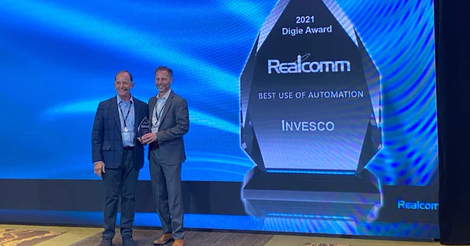 Live event photo from the Realcomm Digie Awards. Two male representatives in business attire from Invesco accept the award on stage from Realcomm for Best Use of Automation.