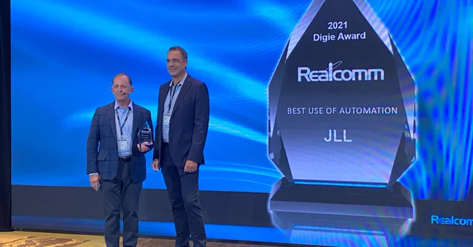Live event photo from the Realcomm Digie Awards. Two male representatives in business attire from JLL accept the award on stage from Realcomm for Best Use of Automation.