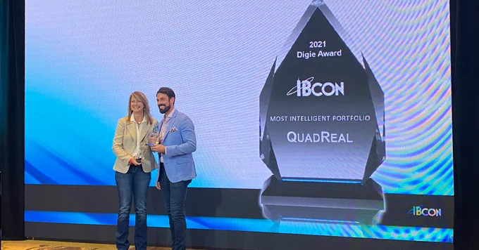 Live event photo from the Realcomm Digie Awards. Two representatives from QualReal, one woman in a tan blazer and one man in a light blue blazer, accept the award on stage from Realcomm for Most Intelligent Portfolio. 
