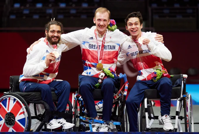 ParalympicsGB men's wheelchair fencing team members Dimitri Coutya, Piers Gulliver and Oliver Lam-Watson pose for a photo while sitting in their wheelchairs. They have their arms around each other, smiling while holding their Paralympic bronze medals from Tokyo 2020.