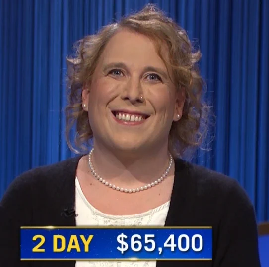 2 Day Jeopardy champ Amy Schneider beams while wearing a black cardigan, white top and a string of pearls. Her hair and makeup are flawless. Text onscreen reads "2 day: $65,400"

