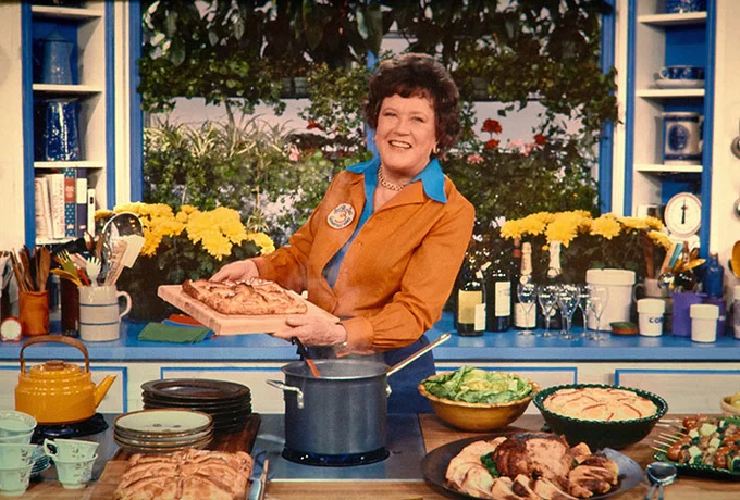 Photo of Julia Child in kitchen, holding bread on a cutting board, surrounded by many courses of a cooked meal and smiling