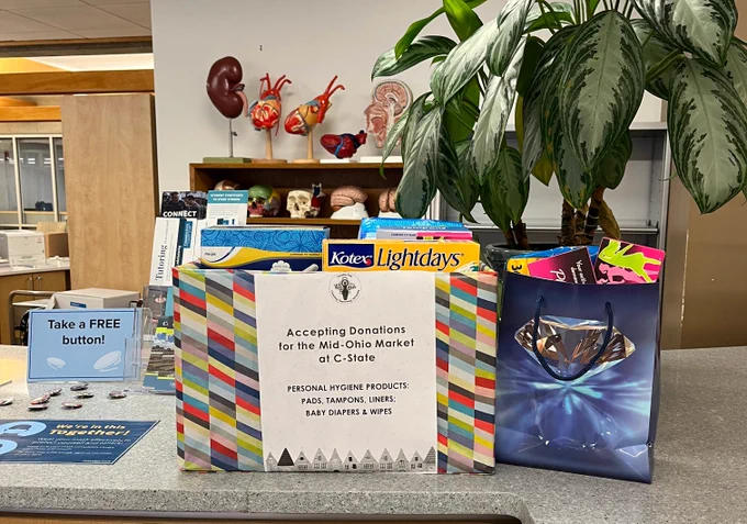 A blue gift bag and a cardboard box covered in wrapping paper sitting on a gray counter. A large plant, anatomical models, and various signage are next to and behind the bag and box. A sign on the box reads: "Accepting Donations for the Mid-Ohio Market at C-State / Personal Hygiene Products: Pads, tampons, liners; baby diapers and wipes."