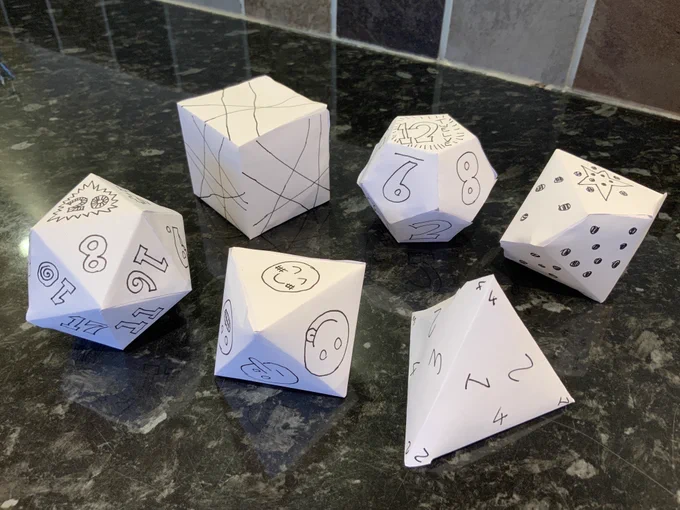 Six paper dice with hand-drawn faces. Each die has a different number of sides, they are labelled with numbers, pips, lines, and one has smiley faces.