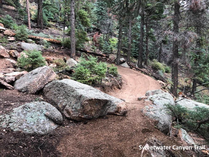 wide, dirt trail winding around large boulders and through a forest.