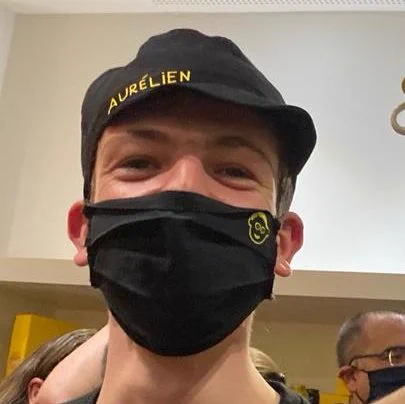 Photographie d'Aurélien. Derrière son masque aux couleurs du café joyeux (noir et jaune), on devine un large sourire. Il porte également une toque de cuisine où est écrit son nom.