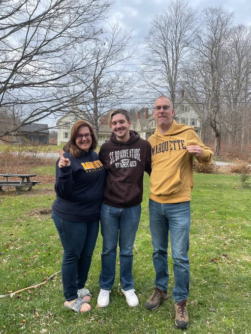 A mother and father wearing Marquette University sweatshirts, standing on either side of their son who is wearing a St. Bonaventure sweatshirt. They are all outside on the front lawn of a house.