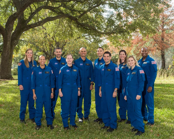 A photo of the 10 members of NASA's new 2021 class of astronaut candidates, each wearing blue flight suits, standing against a fall background of grass and trees