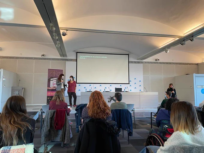 A white conference room and stage with a projector screen, 2 women presenting to a seated audience. To the right of the stage is a sign language interpreter.