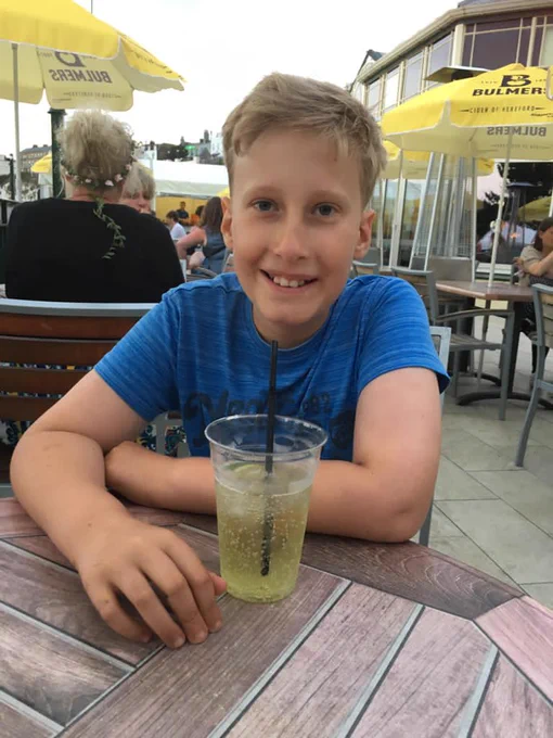 A teenage boy with blonde hair smiles at the camera. He is wearing a blue t-shirt and has a lime and soda with a straw in front of him. He is sitting in a pub beer garden. 