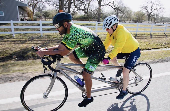Kellie and her pilot are riding the grey Para Guide tandem on a country road; Kellie is wearing a yellow jacket and white helmet; her pilot Anthony is wearing a green and black kit and a black helmet; there is a barn and white fencing in the back ground