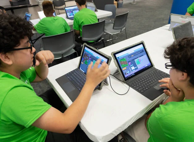 Two students sit at a table working on laptop computers