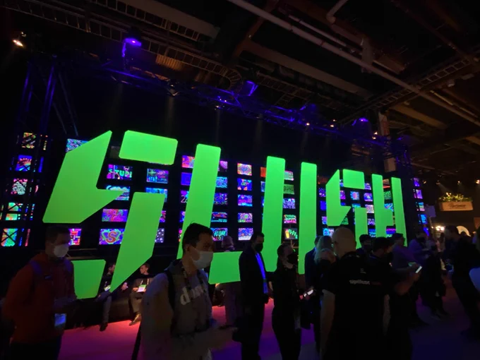 A large neon sign reads "Slush" in capital letters. In front of it, some silhouettes of people are recognizable. They are wearing masks. In the background, many uniform rectangles of neon colors are visible. 