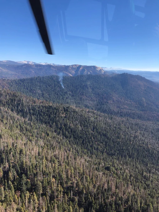 A small plume of smoke rises from a densely forested green mountainside with rugged peaks in the background, seen from above. 