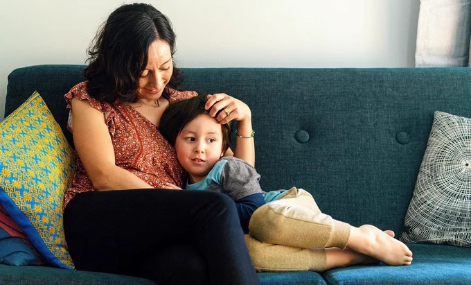 Mother sitting on couch with her arm around her young child, who is curled up next to her.