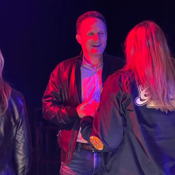 Man laughing, talking to woman whose back is to the camera. Lighting is very red/neon-looking., 