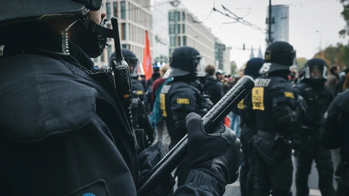 A police officer holding a heavy baton, wearing full gear including helmet, with other police officers in the background, appearing to be watching or preparing to confront protesters.