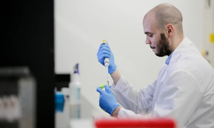 A light-skinned, bald, adult male with a beard wears a white labcoat and blue disposable gloves, as he pipes a solution into a test tube.