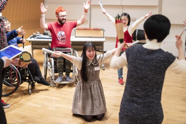 A young Japanese woman kneels in the centre of a circle, smiling broadly with her arms raised aloft. She is holding a percussion instrument and conducting the people around her.
