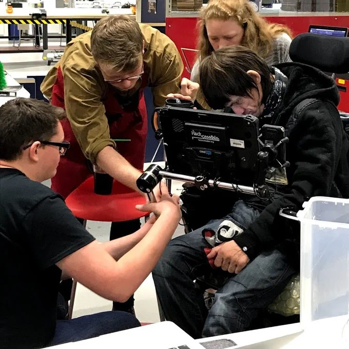 Three members of a team exploring the positioning of a communication aid used by a man pictured sitting down (as part of Hackcessible).