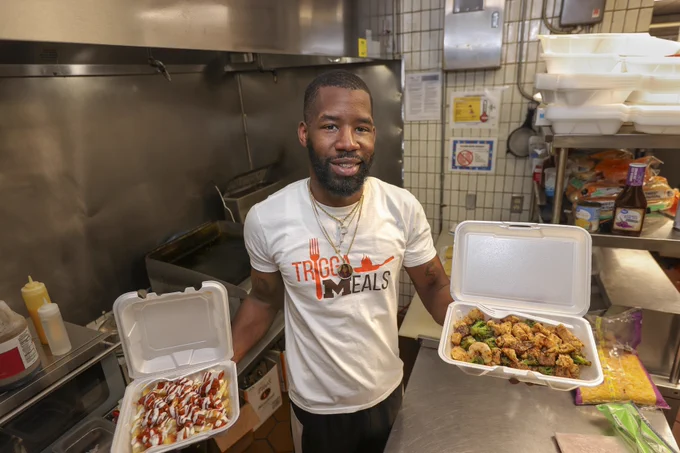 Deante Shivers holds two plates of food in each hand, standing in the kitchen