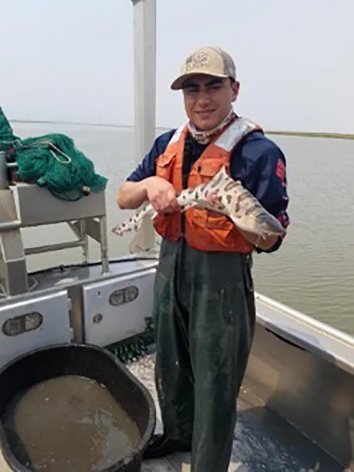 A person in a khaki hat, orange vest, and dark green pants holding a spotted sea creature while standing on a boat.