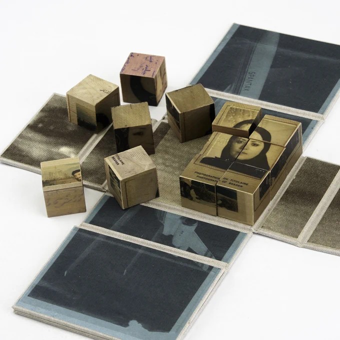 Detail of an unfolded artist’s book from a three-quarter perspective, printed with a blue airplane and a sepia eye. Twelve wooden blocks rest on top. Six are scattered, and the other six are aligned in a grid, depicting a woman in a black headscarf.