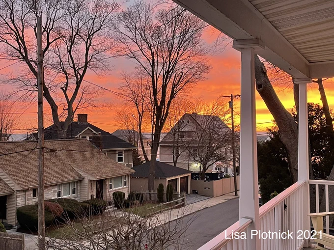 Sunrise as viewed from a front porch. An orange glow is on the horizon to the far right, adding a pinkish hue to the clouds to the left. Houses and tall bare trees line the street on the left side.