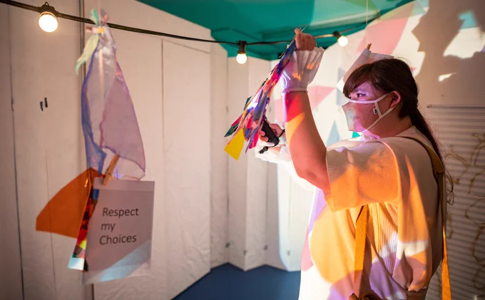 A woman with brown hair, a clear face mask and white costume hangs a colourful item on a suspended wire. Another piece of card hanging from the wire says 'respect my choices'.