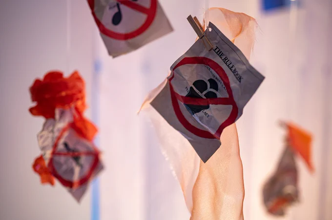 Colourful pieces of paper hang from the ceiling. The image in the foreground shows a clear image saying 'the rulebook' and with a picture of a cake overlain with a red line.