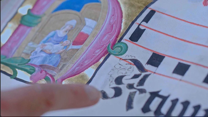 A white woman’s finger points to a closeup of large black gothic script below music notes. To the left is an illuminated letter with pink and gold paint depicting Saint Anne cradling the Virgin Mary.