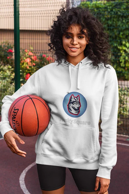 A smiling young adult woman, is wearing a beige 'Husky Square' hoodie sweatshirt with a basketball tucked between her right arm and right hip