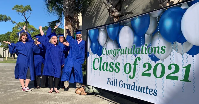 A group of celebrating grads in caps and gowns stand next to a banner that reads, "Congratulations Class of 2021 Fall Graduates."