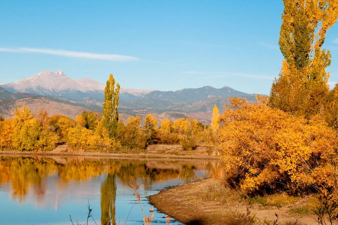 Photo of Longmont Colorado in fall. A calm river and orange-leafed trees paint the foreground, a large mountain range is framed in the background.