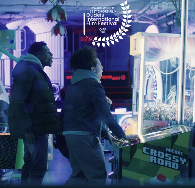 Two young men celebrate as they play a video game in a dark arcade. Winning film festival laurel top middle of the frame